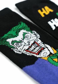 Calzini neri con una grafica colorata di un personaggio con capelli verdi, viso bianco e labbra rosse, il testo recita "HA HA" in giallo e bianco.