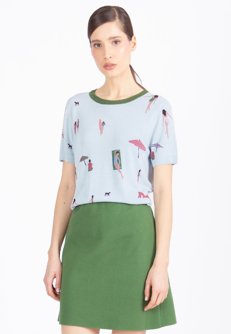 Jasnoniebieski sweter z krótkim rękawem z kolorowymi, plażowymi wzorami, zestawiony z zieloną, dopasowaną spódnicą. Miękki, dzianinowy materiał i zaokrąglony dekolt.