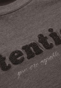Grå sweatshirt med texturerad svart bokstavering som står "uppmärksam" och en diskret vit tryckning som säger "du är kapabel."