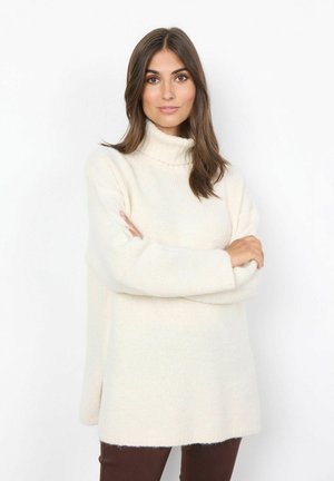 Cremfarbener Rollkragenpullover mit lockerer Passform, grober Strickstruktur, langen Ärmeln und seitlichen Schlitzen. Kombiniert mit braunen, eng anliegenden Hosen.