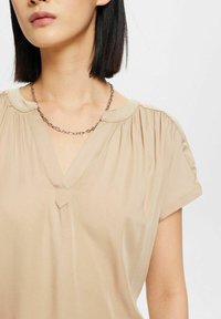 Blouse beige à manches courtes avec un col en V et des fronces aux épaules. Présente un petit détail triangulaire au niveau du décolleté et une texture lisse.