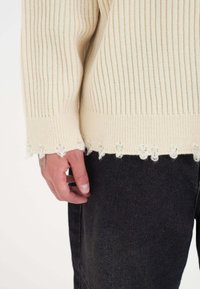 Beige gestreifter Pullover mit ausgefranstem Saumdetail. Verfügt über lange Ärmel und strukturiertes Material. Kombiniert mit dunklen Jeans, die einen lässigen Stil zeigen.