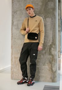 Sudadera naranja, pantalones cargo negros, bolso peludo negro y zapatillas con acentos rojos. El modelo lleva un gorro naranja frente a un fondo de hormigón.