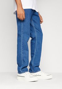 Vaqueros de mezclilla azul con corte recto y costuras laterales visibles, combinados con zapatillas blancas, en primer plano sobre un fondo neutro.