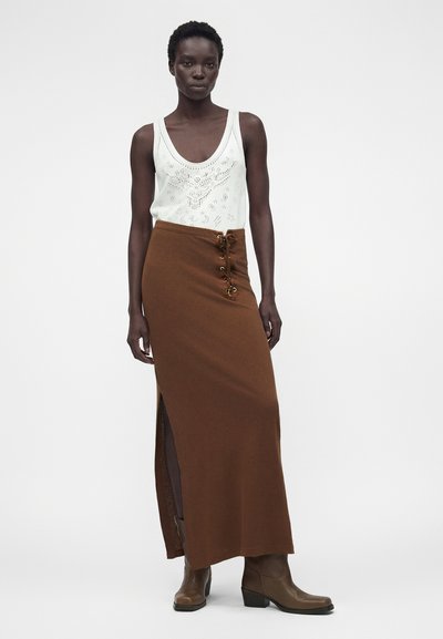 Μαύρη γυναίκα που φοράει λευκό αμάνικο τοπ με λεπτομέρειες από τρυπητό ύφασμα, μακριά καφέ φούστα με ψηλή μέση, σκίσιμο στο πλάι και λεπτομέρειες δέσιμου, και καφέ μπότες.