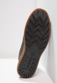 Suela de zapato de goma negra con un patrón en zigzag y agujeros de ventilación, con un borde marrón. Marca Geox destacada en el centro.
