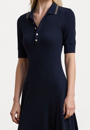 Femme portant une robe polo bleu marine à manches courtes avec un col orné de bordures dorées et trois boutons dorés sur la patte de boutonnage avant.