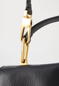 Sac à main en cuir texturé noir avec un fermoir en métal doré brillant et une sangle en cuir noir. Gros plan sur le détail d'attache.