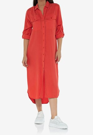 Mujer con vestido camisero rojo de manga larga con las mangas remangadas y zapatillas blancas, de pie sobre un fondo blanco.