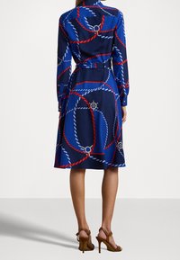 Námořnické šaty s vzorem červené a bílé šňůry, dlouhými rukávy, páskem v pase a siluetou ve tvaru A. Obsahují zapínací manžety a dekorativní detaily.