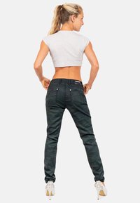 Donna con coda di cavallo bionda, che indossa un crop top bianco, pantaloni mimetici scuri e tacchi bianchi con punte, in piedi con le mani sui fianchi, di spalle.