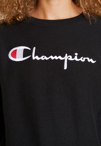 Gros plan d'un sweat-shirt noir avec un logo Champion blanc et rouge brodé sur la poitrine, porté par une personne aux cheveux bouclés.