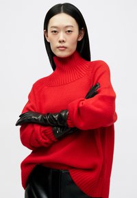 Red knit sweater with a high ribbed collar and loose fit, paired with black leather gloves, shot against a light background.