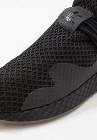 Svart idrottssko med en strukturerad mesh-övre, släta svarta accenter och en gummisula. Logotypen visas på toppen.