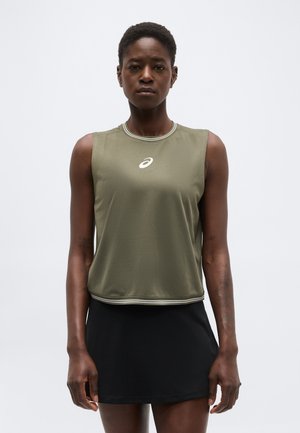 Atletische vrouw die een mouwloze olijfgroene top met logo en een zwarte rok draagt, staand tegen een effen lichte achtergrond.