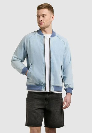 Junger Mann, der eine hellblaue Bomberjacke mit Reißverschluss, ein weißes T-Shirt und schwarze Jeansshorts trägt, steht mit einer Hand in der Tasche.