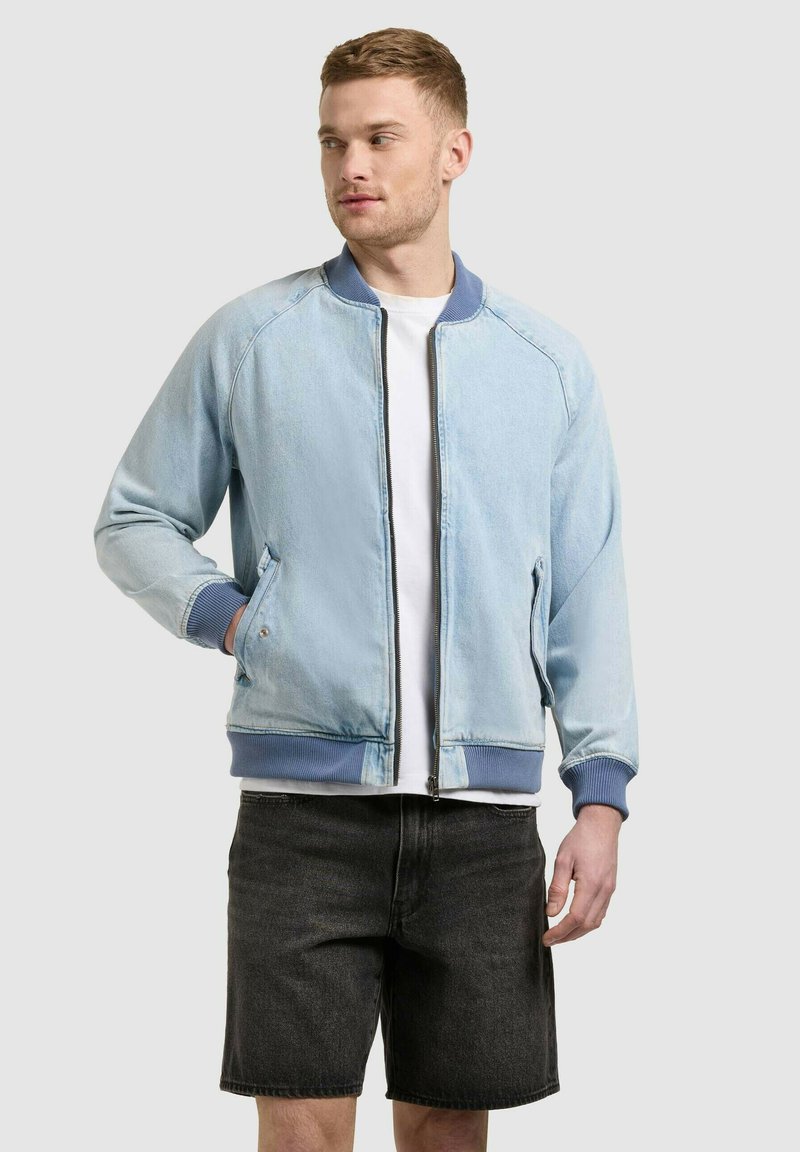 Jeune homme portant une veste bomber zippée bleu clair, un t-shirt blanc et un short en jean noir, debout avec une main dans la poche.