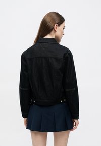 Melna džinsa jaka ar strukturētu dizainu, redzamām šuvēm un pogu detaļām viduklī. Valkāta pār tumši zilu plisētu svārku.