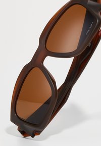 Brune solbriller med mat overflade, vinklede rammer og tonede linser. Designet har stærke linjer og en bred stang for stabilitet.