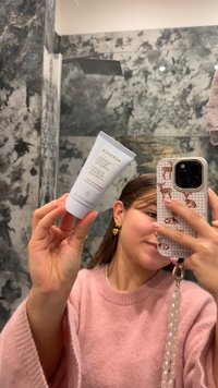 Tube der Kérastase-Haarbehandlung, weiß mit grauem Text, in einer Hand gehalten. Texturierter rosa Pullover sichtbar. Dekorative Handyhülle mit Tiermuster.