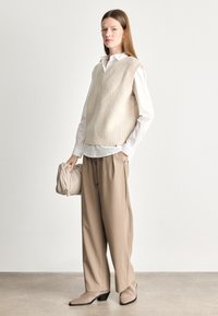 Colete de malha bege com decote em V, usado por cima de uma camisa branca, combinado com umas calças largas bege e uns sapatos de salto agudo bege. O modelo segura uma bolsa texturizada bege.