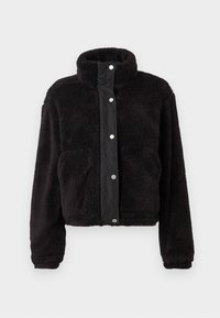 Svart, fluffig fleecejacka med ståkrage, framknäppning med tryckknappar, långa ärmar och två sidofickor mot en ljus, enkel bakgrund.