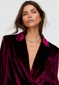 Samtblazer in tiefem Burgunderrot mit Satin-Kragen, Einzelknopfverschluss und glatter Textur. Auffällig durch das tiefe V-Ausschnitt-Design.