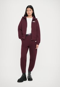 Burgundy kapucnis pulóver és joggingnadrág, puha anyagból, cipzáros elülső résszel, oldalsó zsebekkel és logós derékpánttal. Fekete bokacsizmák.