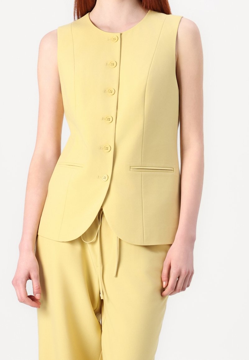 Femme portant un gilet jaune sans manches à boutons avec deux poches frontales et un pantalon ample assorti sur fond blanc.