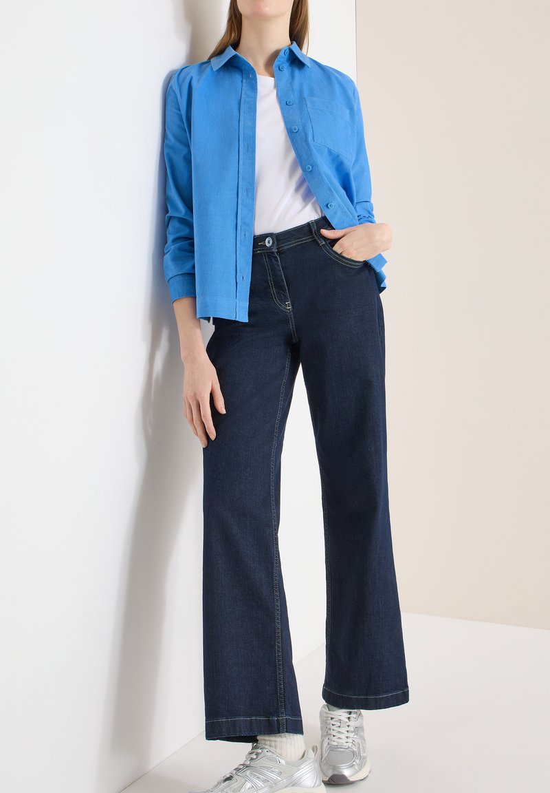 Femme appuyée contre un mur, vêtue d'une chemise bleue boutonnée par-dessus un haut blanc, d'un jean large foncé, de chaussettes blanches et de baskets argentées.