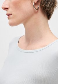 Camisa cinza claro com textura canelada e decote redondo. A pessoa usa um brinco de argola prateado, destacando a orelha e o perfil do rosto.