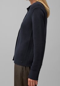 Marinblå stickad kavaj med strukturerat mönster, knappstängning och långa ärmar. Har en slät tygkvalitet och ribbade ärmslut.