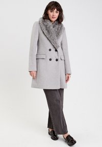 Abrigo gris de doble botonadura con collar de piel sintética, que presenta dos bolsillos frontales y botones negros, llevado sobre pantalones de pierna ancha en gris oscuro.