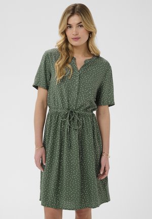 Nő zöld, rövid ujjú ruhát visel fehér pöttyökkel, elöl gombokkal és megkötős derékkal, egy világos, egyszerű háttér előtt áll.