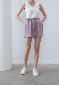 Haut blanc sans manches avec des accents à volants et short lavande avec taille à nouer. Le mannequin porte des baskets blanches sur un sol en béton.