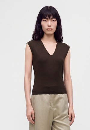 Femme aux longs cheveux foncés portant un haut sans manches marron foncé à col en V et un pantalon beige, debout devant un fond clair uni.