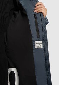 Puffy bunda v tmavosivej farbe s čiernym interiérom. Obsahuje bočný zips na vrecko a nálepku s nápisom "Teplé objatie vyrobené z látky."