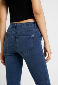 Tummansiniset denimi-farkut slim-fit-mallilla, joissa on takataskut, ei näkyviä kulutuksen merkkejä ja sileä pinta. Vyötärönauha istuu juuri lantiota ylempänä.