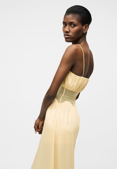 Γυναίκα με σκουρόχρωμο δέρμα και μαλλιά, ντυμένη με κίτρινο αμάνικο φόρεμα με λεπτές τιράντες, κοιτάζοντας πάνω από τον ώμο της σε ένα απλό φόντο.