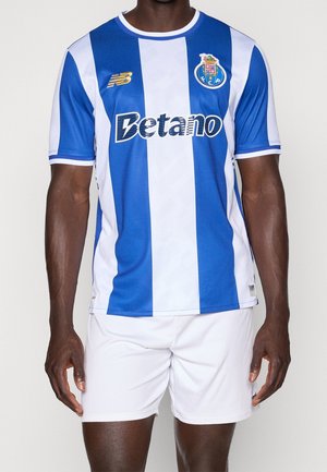 Mann trägt blau-weiß vertikal gestreiftes FC Porto Fußballtrikot mit Betano-Sponsor und weißen Shorts, stehend vor einfarbigem Hintergrund.