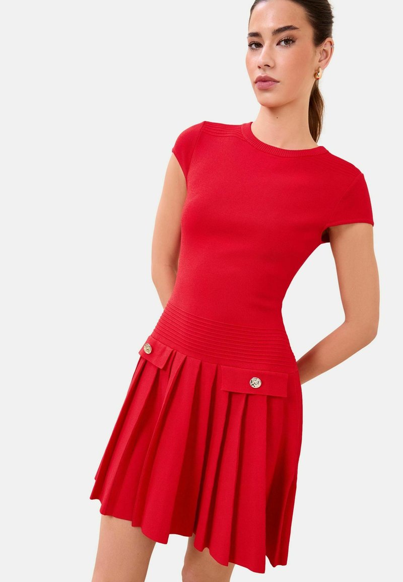 Sieviete valkā piegulošu sarkanu īsrokas kleitu ar ribotu jostas daļu un pieklājīgu svārku, kurā ir divas priekšpuses atloku kabatas ar pogām.