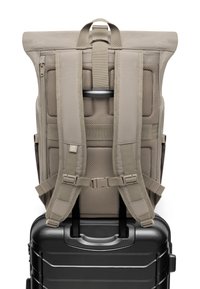 Zaino con spallacci imbottiti in beige chiaro, dotato di apertura a roll-top e tessuto a rete per la ventilazione, appoggiato su una valigia rigida nera.