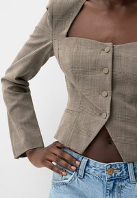 Hnědý károvaný blejzr se čtvercovým výstřihem, zapínáním na knoflíky a dlouhými rukávy, doplněný světle modrými džínami s vysokým pasem.