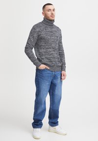 Dunkelgrauer, strukturierter Rollkragenpullover mit langen Ärmeln, kombiniert mit hellblauen Jeans und weißen Sneakers mit gelben Akzenten.