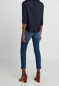 Marinblå skjorta med knappar och trekvartsärmar, kombinerad med åtsittande blå jeans och bruna remsandaler, sedd bakifrån.