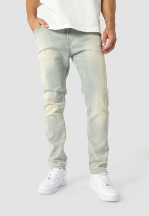 Mann trägt hellblaue Slim-Fit-Jeans, weiße Sneakers und ein weißes Hemd, steht mit einer Hand in der Tasche vor einem einfarbigen Hintergrund.