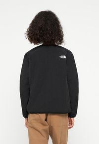 Personne aux cheveux bouclés portant une veste noire North Face et un pantalon beige, debout dos à la caméra sur un fond uni.