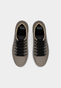 Ζευγάρι ταμπά casual αθλητικά παπούτσια με μαύρα κορδόνια, μπεζ περίγραμμα και λευκές σόλες, απεικονιζόμενα από πάνω σε λευκό φόντο.