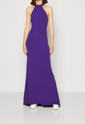 Kvinde iført en ærmeløs, gulvlængde lilla kjole med halterneck og hvide plateau-sko, stående foran en ensfarvet baggrund.