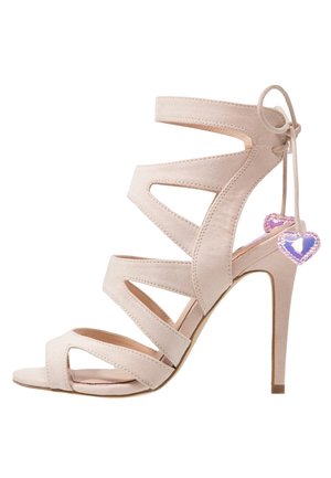 Sandale à talons hauts en daim beige avec plusieurs sangles découpées et un lien de cheville, ornée de deux petits charms en forme de cœur violet.